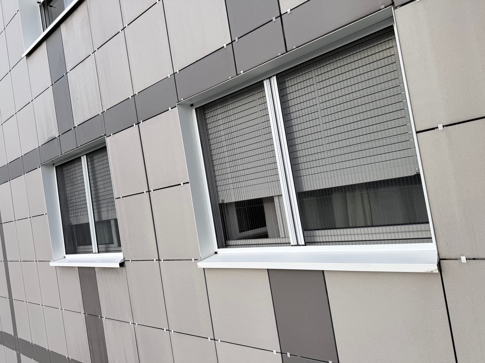 Hausfassade von aussen mit zwei Fenstern, an denen das Insektenschutz-Plissee montiert ist. Der helle Rahmen sitzt vor dem Fensterrahmen und zeichnet sich deutlich ab.