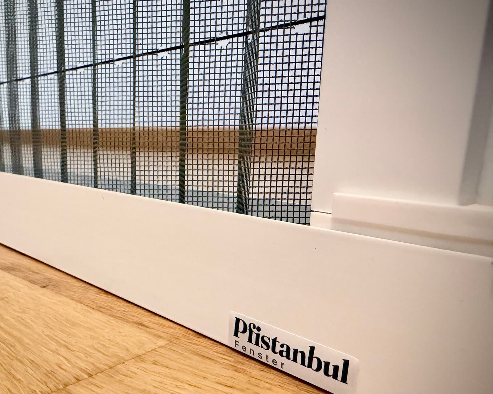 Die untere Schiene eines Insektenschutz-Plissees auf einem Eichenparkett, darüber das graue Gewebe. Auf der Schiene klebt ein Aufkleber mit dem Schriftzug Pfistanbul Fenster.